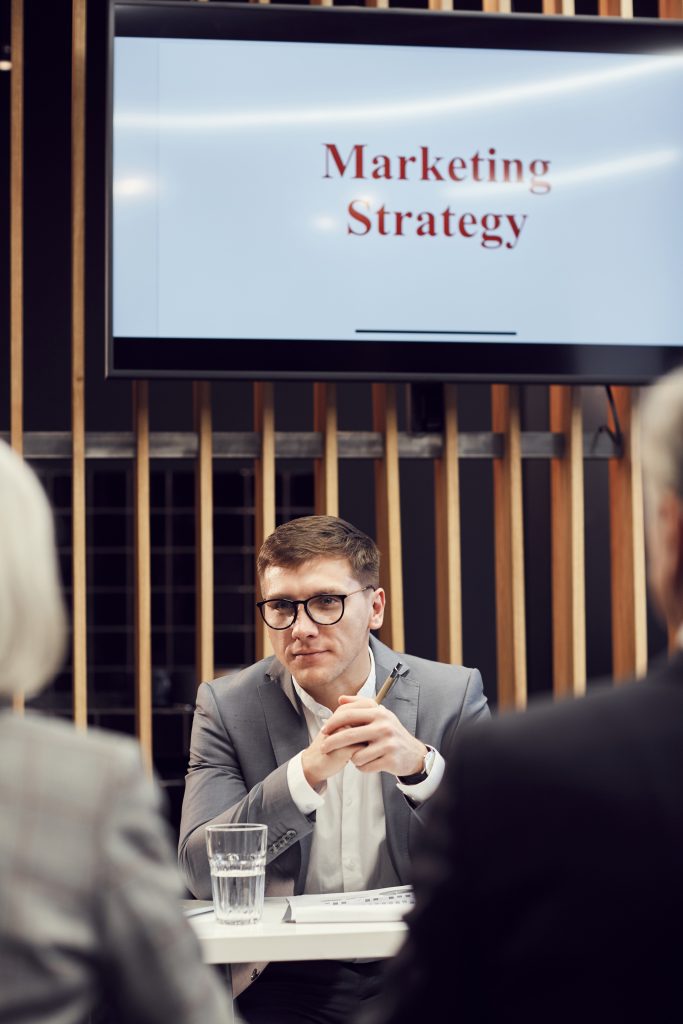 Content experienced young marketer in eyeglasses sitting at table with glass of water and talking to entrepreneurs at conference