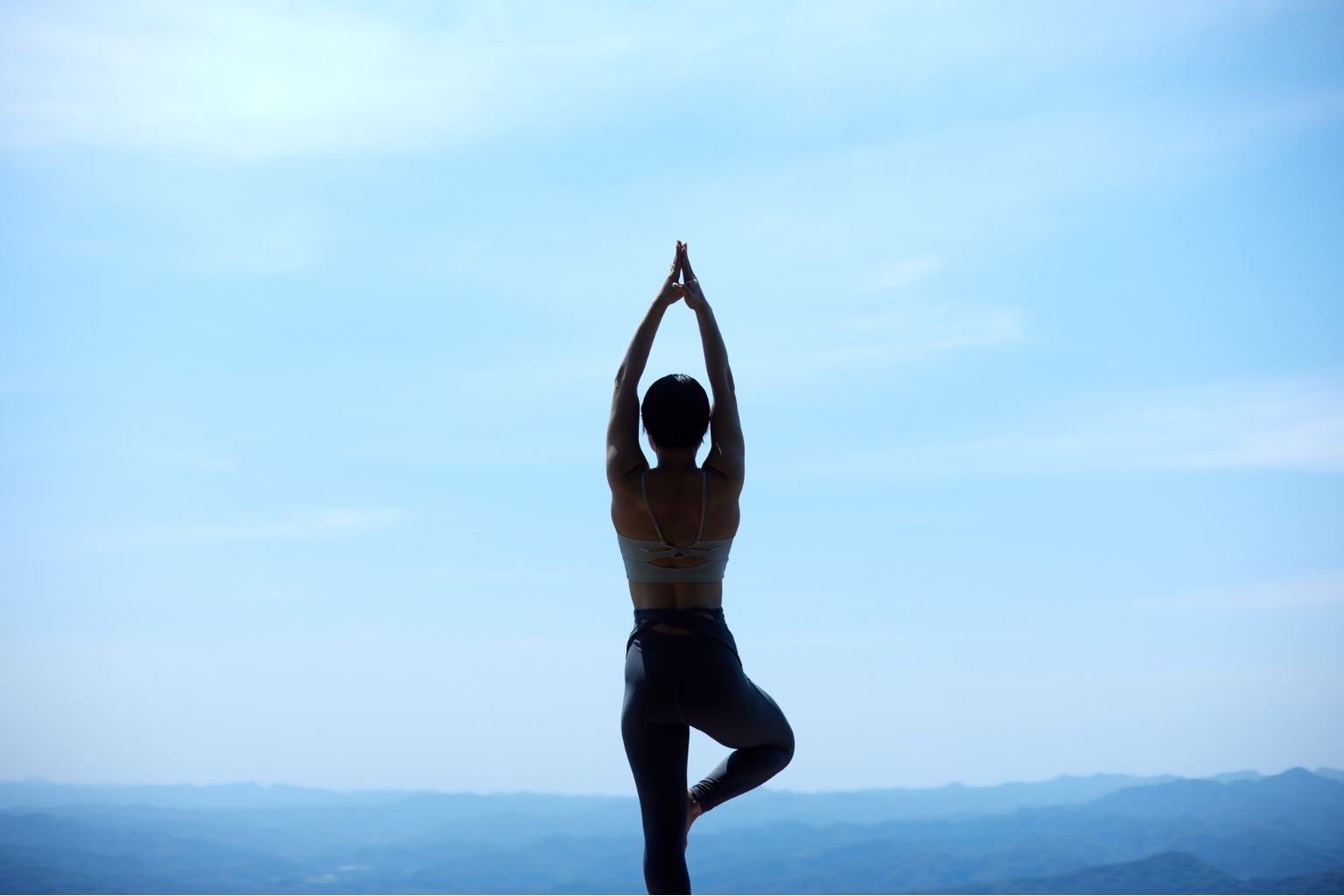 Silhouette of a person practicing yoga in tree pose against a serene blue sky and distant mountain landscape.